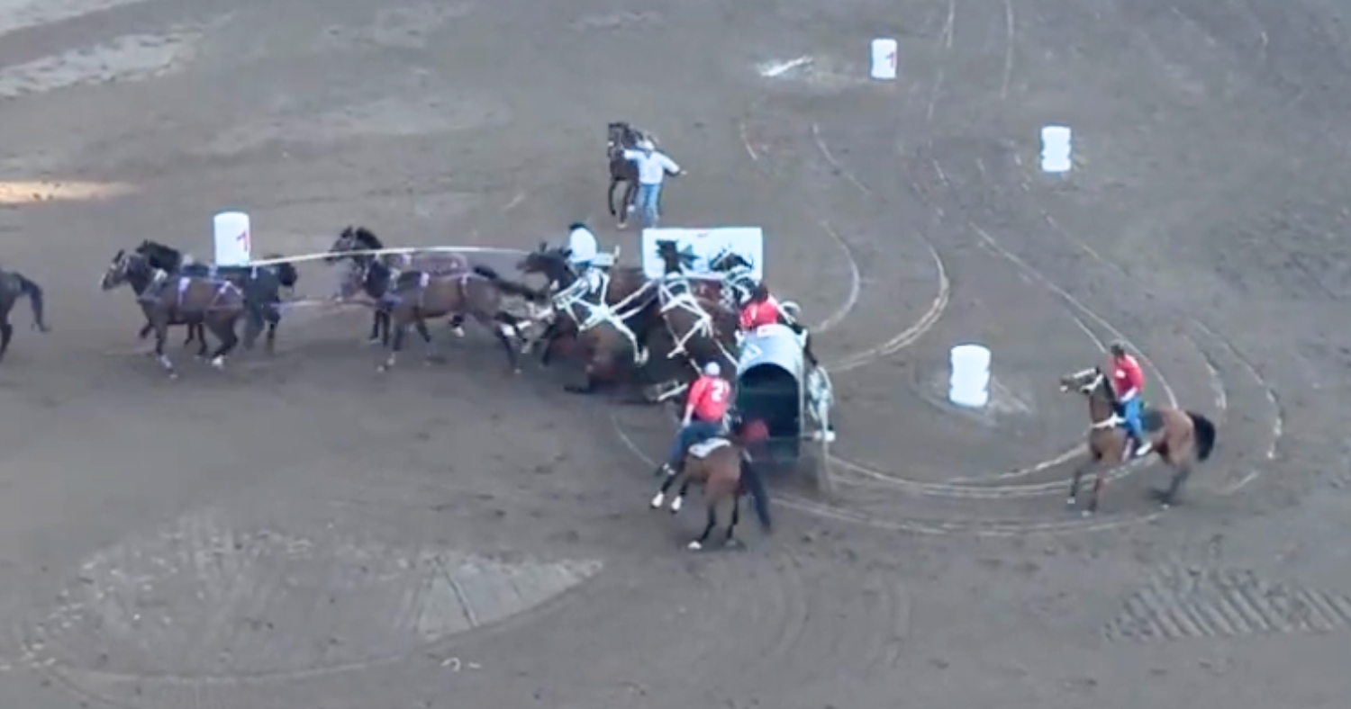 Overhead view of a dramatic chuckwagon race collision on a dirt track. Multiple dark-colored horses hooked to wagons are caught in a pile-up, with a large, bright red circle drawn around the center of the crash to highlight the entangled horses.